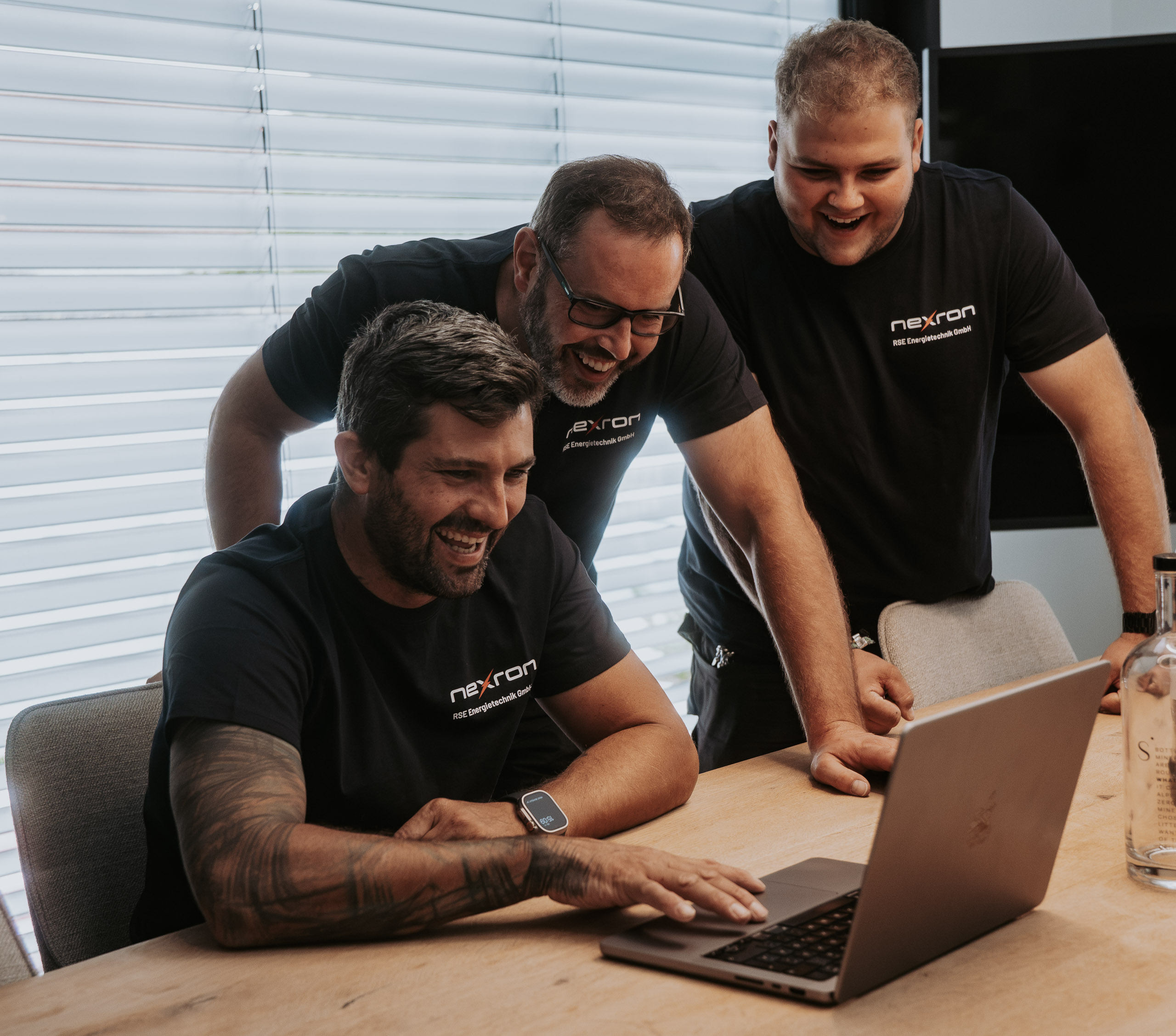 Drei lächelnde Männer in schwarzen T-Shirts der Firma nexron RSE Energietechnik GmbH schauen gemeinsam auf einen Laptop-Bildschirm an einem Holztisch.