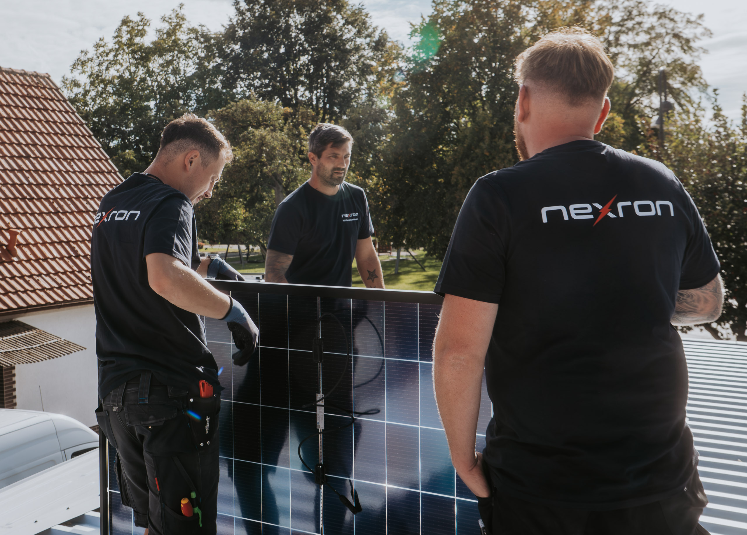 Drei Handwerker der Firma Nexron Energietechnik heben und montieren gemeinsam ein Solarmodul auf einem Dach.