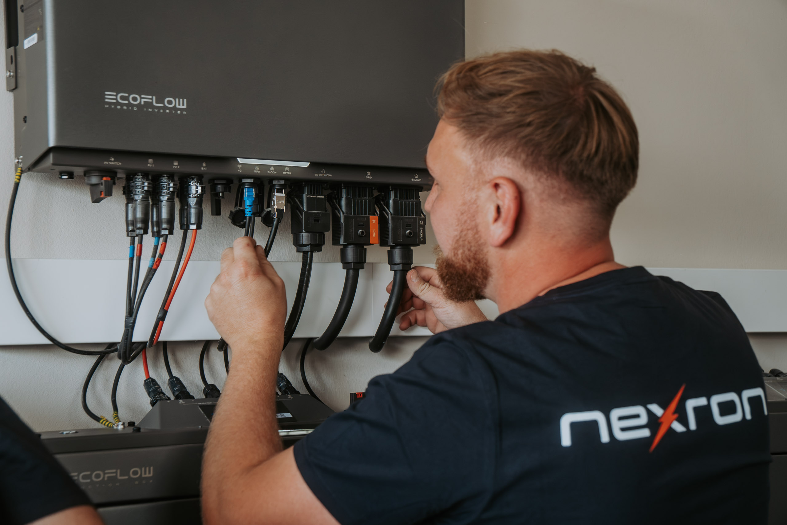 Ein Techniker im Nexron-Shirt schließt präzise Kabel und Anschlüsse an der Unterseite eines wandmontierten EcoFlow Hybrid-Wechselrichters an.