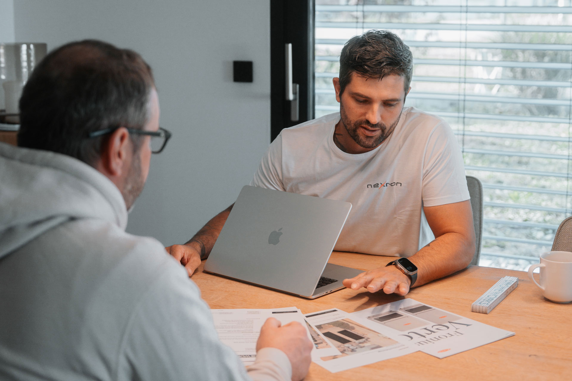 Ein Nexron-Experte im weißen Marken-T-Shirt erklärt einem Kunden an einem Holztisch im Detail die Planung eines Photovoltaik-Systems anhand von Datenblättern und einem Laptop.